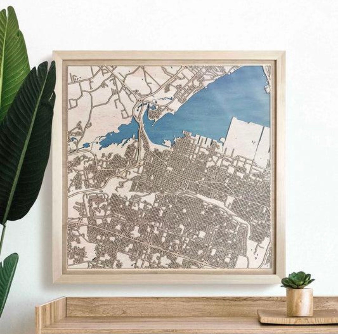 A framed wooden city map displayed on a console table with plants